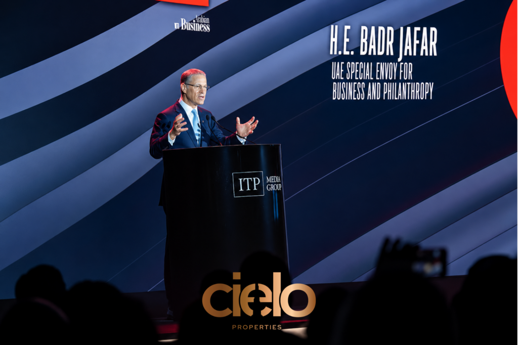 Speaker stands at a podium addressing an audience at a business conference. He is wearing a dark suit and gesturing with both hands while speaking. Behind him, a large illuminated stage screen displays the text “H.E. Badr Jafar, UAE Special Envoy for Business and Philanthropy,” along with Arabian Business and ITP Media Group branding. The backdrop features diagonal blue and gray graphics, while audience silhouettes are visible in the foreground.