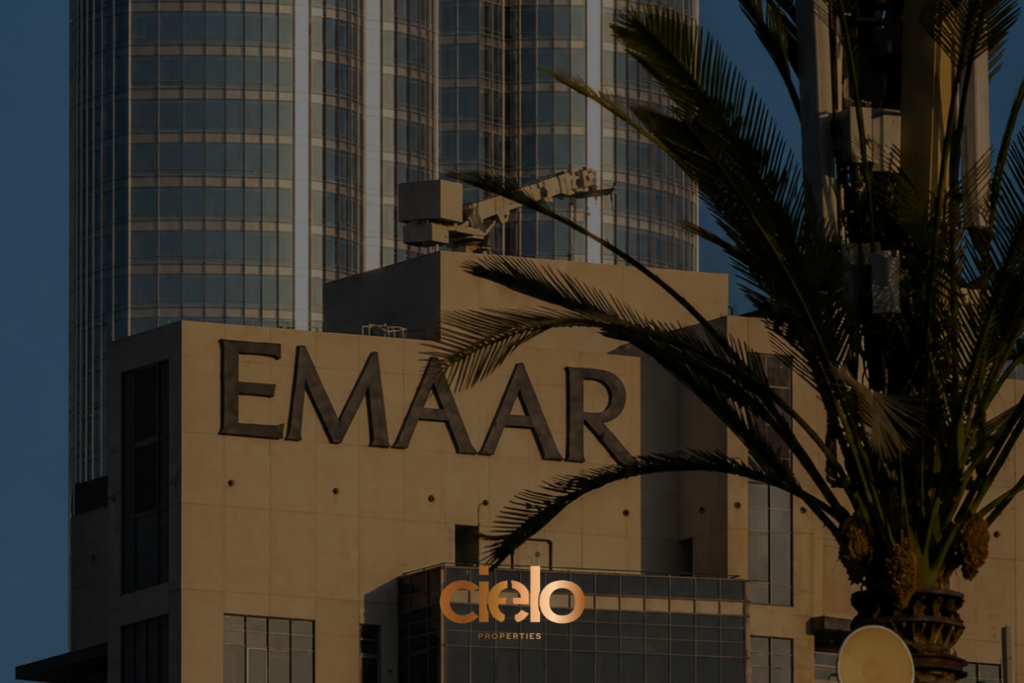 **Alt text:** *Exterior view of an Emaar building in warm golden-hour light, featuring the large “EMAAR” sign on a beige facade. Glass skyscraper towers rise in the background, while palm fronds partially frame and obscure the scene.