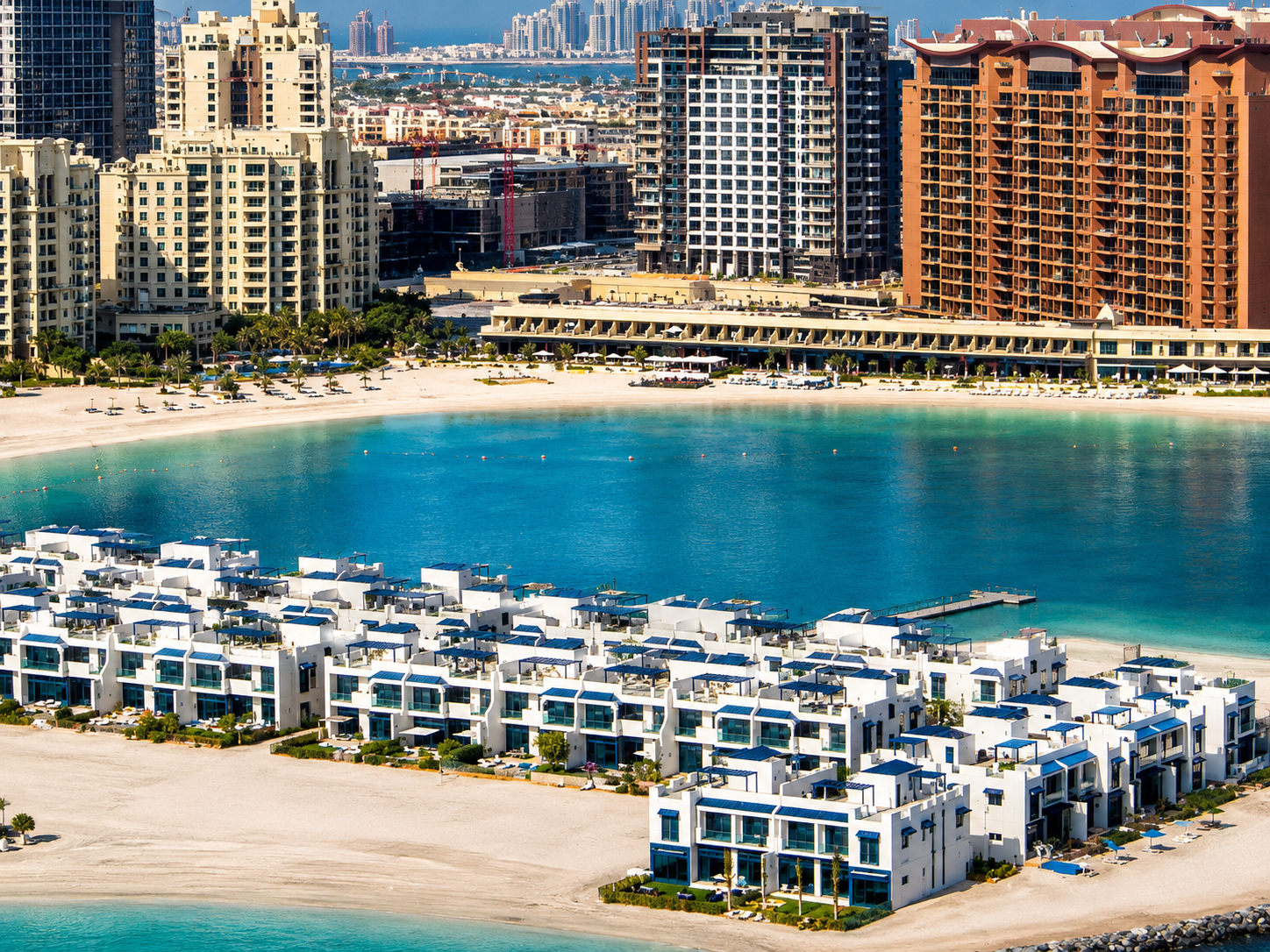 A panoramic waterfront view of Palm Jumeirah in Dubai, featuring a row of modern white beachfront villas overlooking a calm turquoise lagoon. Behind the shoreline, luxury residential towers and hotels rise against the skyline, with Palm Jumeirah's iconic urban landscape visible in the distance. The scene highlights pristine sandy beaches, crystal-clear waters, and upscale coastal living under a bright blue sky.