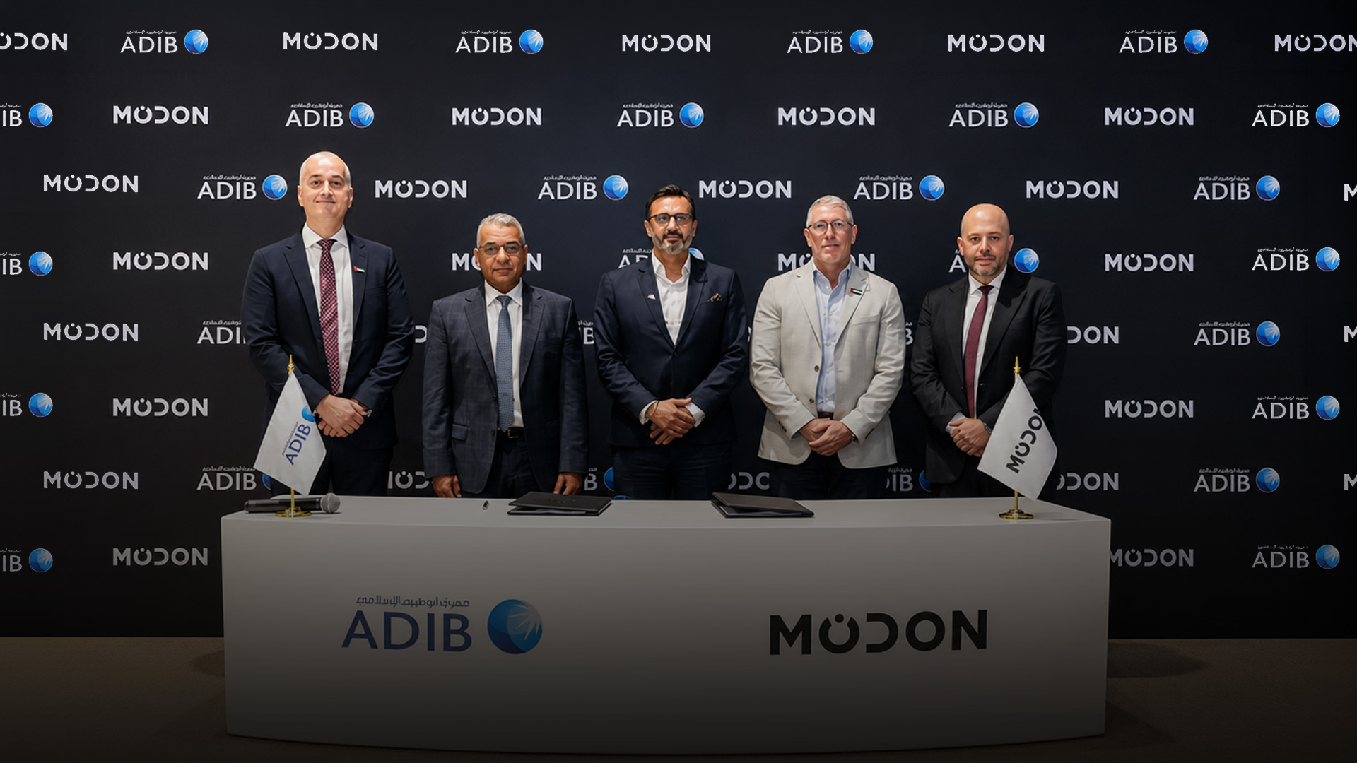 **Alt text:** A group of five executives stands behind a signing table at a formal partnership event between Modon and Abu Dhabi Islamic Bank (ADIB). The backdrop features repeating Modon and ADIB logos, while branded flags and signed documents are displayed on the table, symbolizing the announcement of a strategic collaboration to introduce Abu Dhabi's first off-plan home financing solutions.